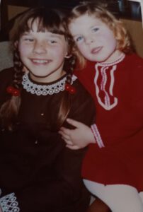 Zwei Mädchen sitzen auf einem Sessel und lächeln in die Kamera. Die kleine Blonde trägt ein rotes Kleid. Die dunkelhaarige Schwester trägt ein braunes Kleid. Das Bild ist ungefähr 1969 entstanden.