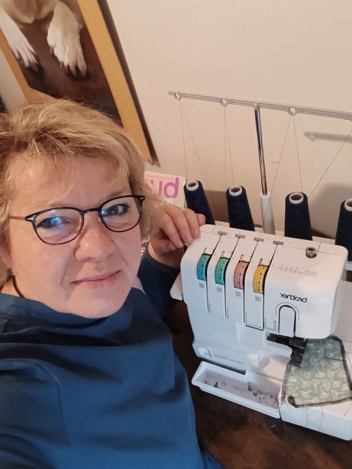 Frau mittleren Alters sitzt vor einer Overlocknähmaschine und schaut in die Kamera