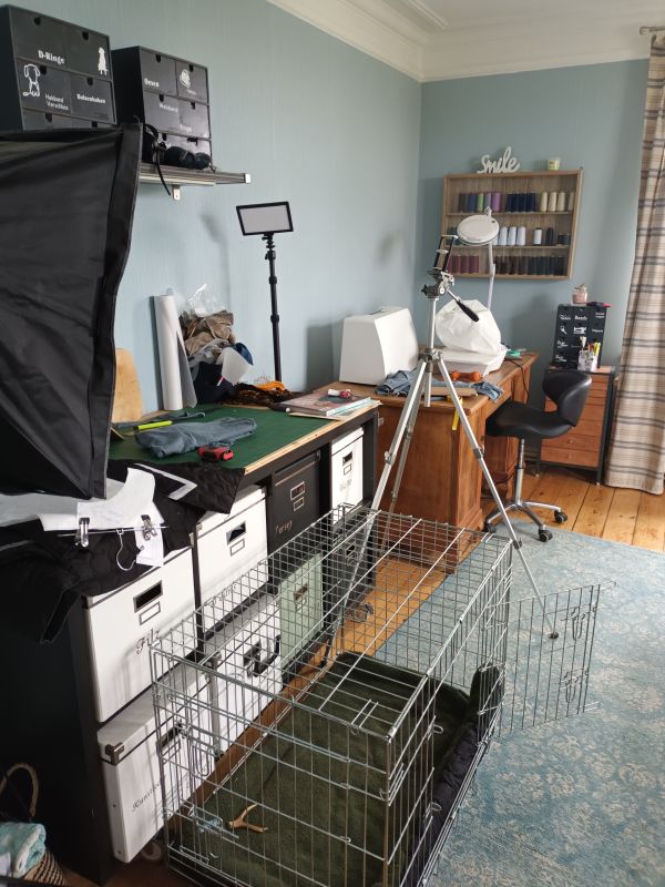 Es ist ein Zimmer zu sehen in dem ziemlich viel Chaos herrscht. Es ist ein Nähzimmer, das dringend Struktur benötigt. Im Vordergrund steht ein Hundekennel - ohne Hund.